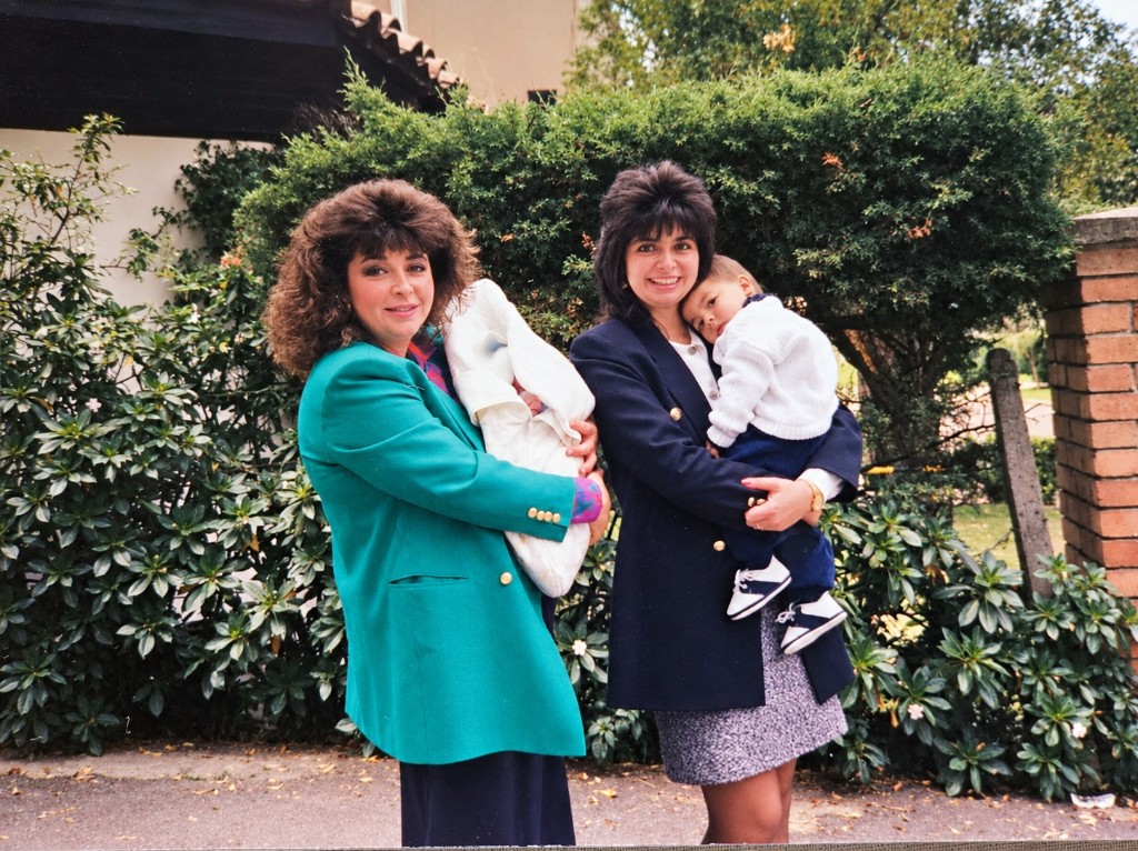 Las dos hermanas con sus bebés en brazos, frente a un seto, el día del bautizo de Nico